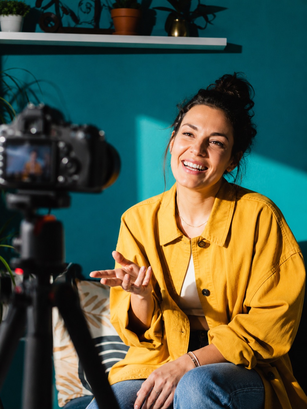 Une femme en veste jaune parle face à une caméra sur trépied, devant un mur turquoise