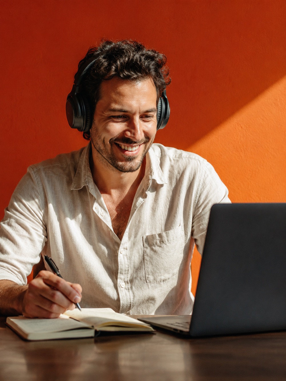 Un homme au casque prend des notes devant un ordinateur ouvert, devant un mur orange